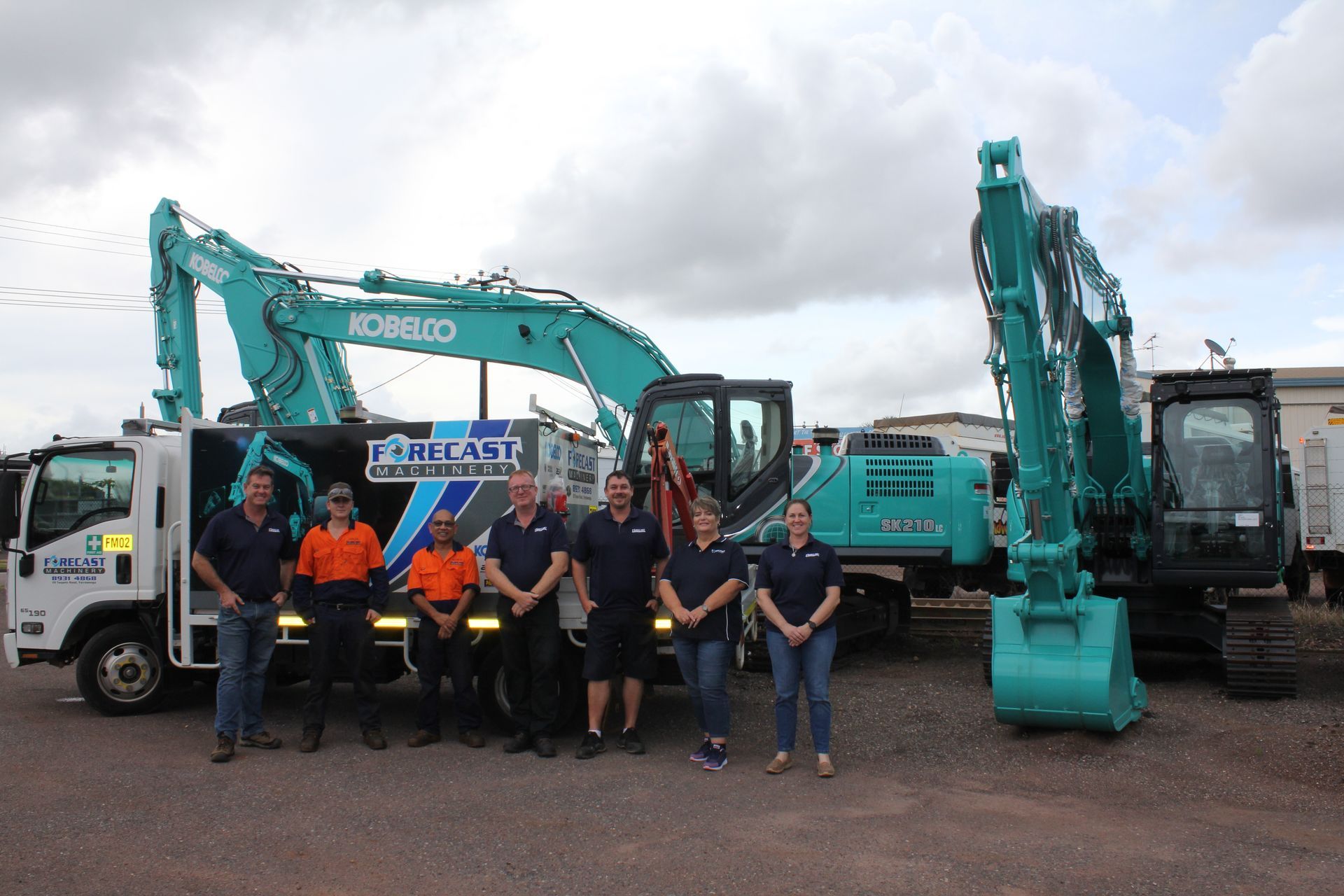 Six people stand in a row in front of a white service truck and two teal Kobelco excavators at an outdoor site — Forecast Machinery in Yarrawonga, NT