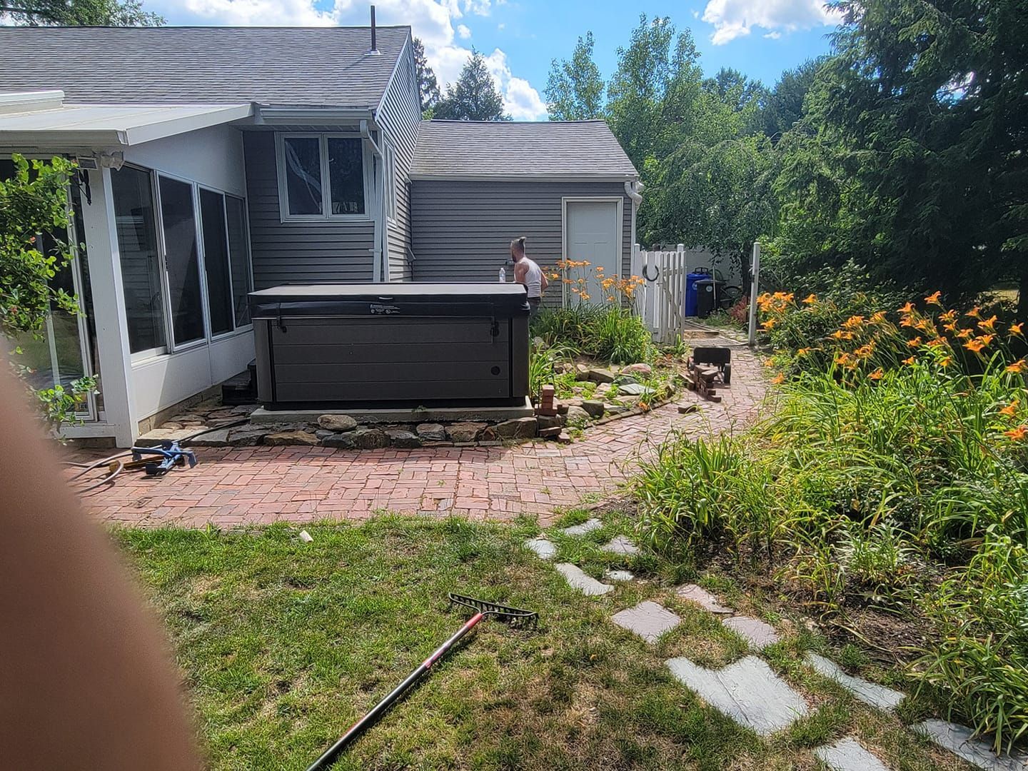 A black hot tub sits on a brick patio beside a house with a person in the background. Sunny outdoor setting.