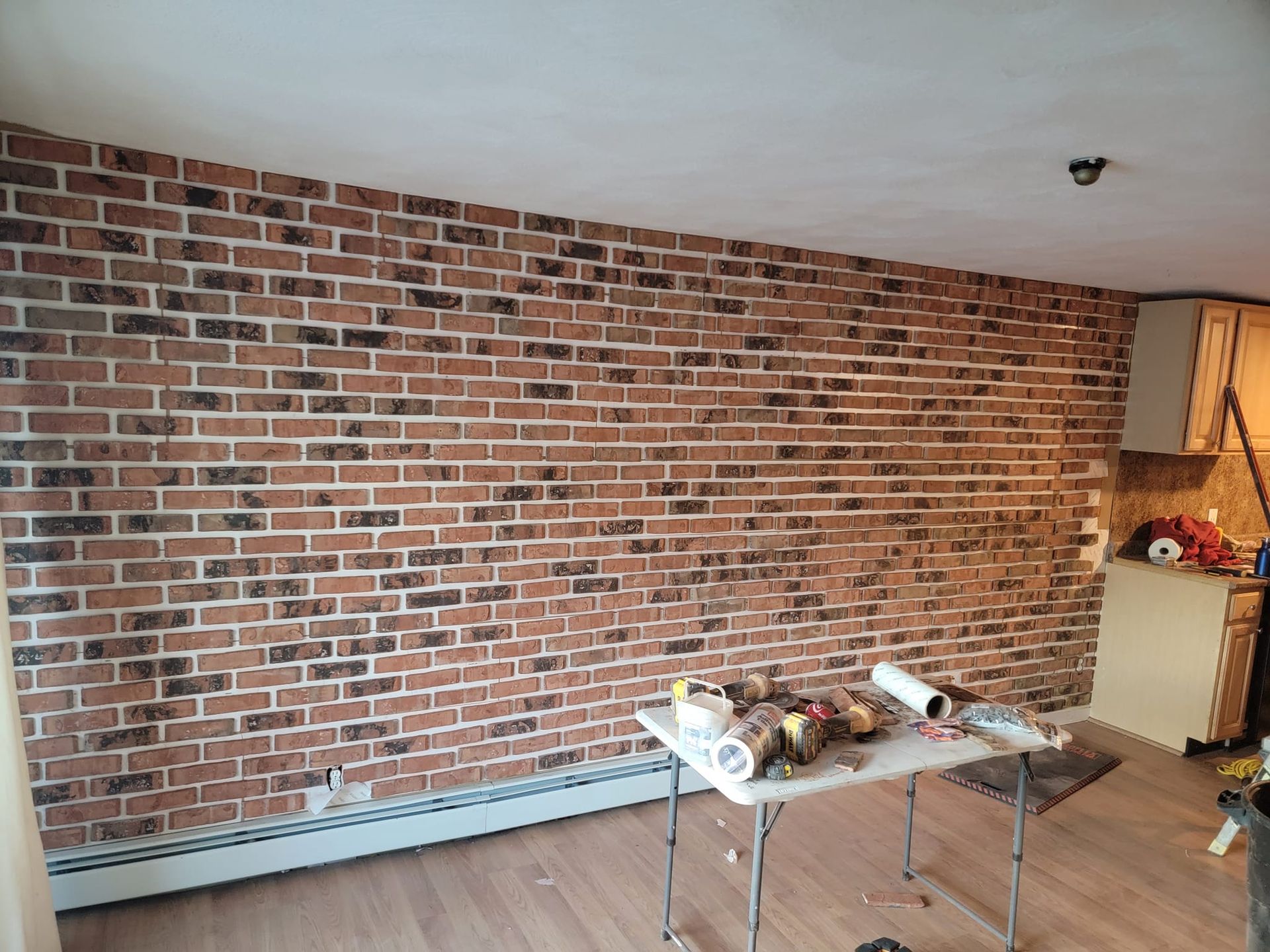 Brick veneer wall in a room, with a folding table holding supplies. Light wood flooring and white ceiling.