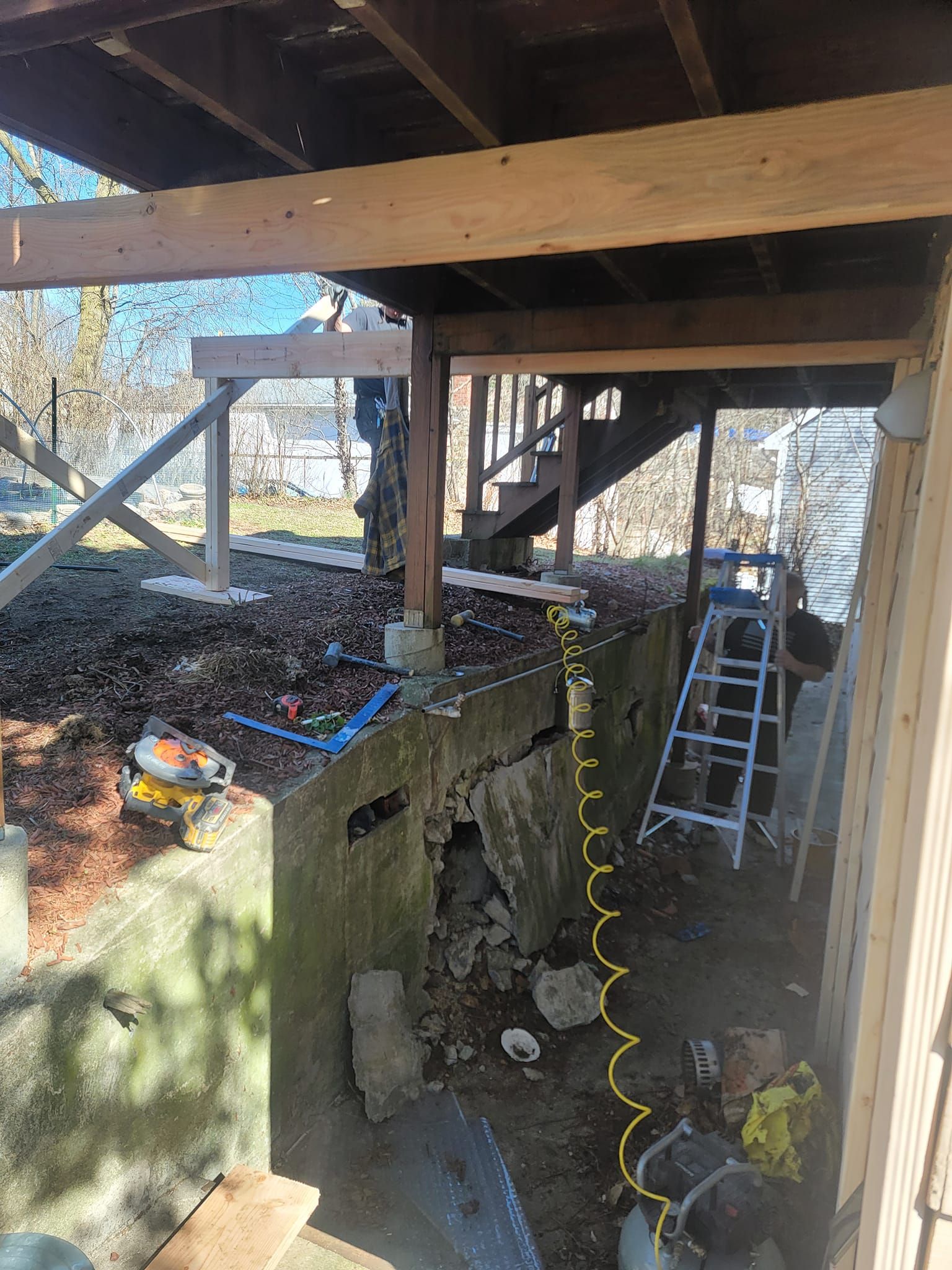 Construction site under a deck with exposed concrete foundation, stair access, and tools.