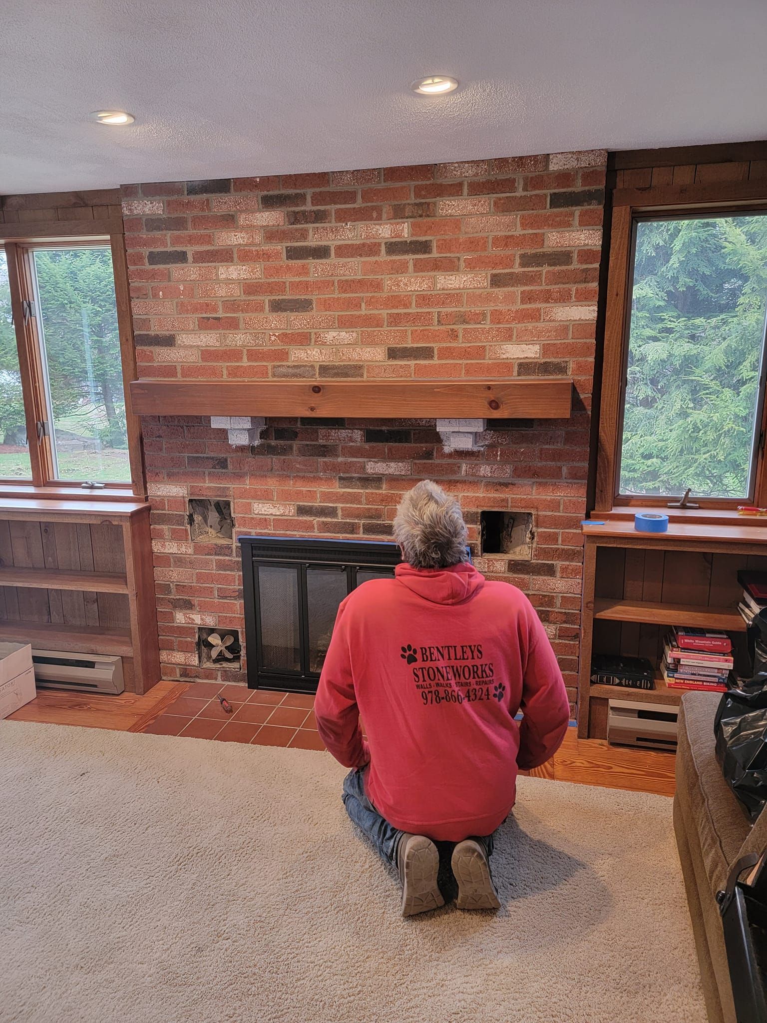 Person kneeling in front of a brick fireplace with a wooden mantel, between two windows.
