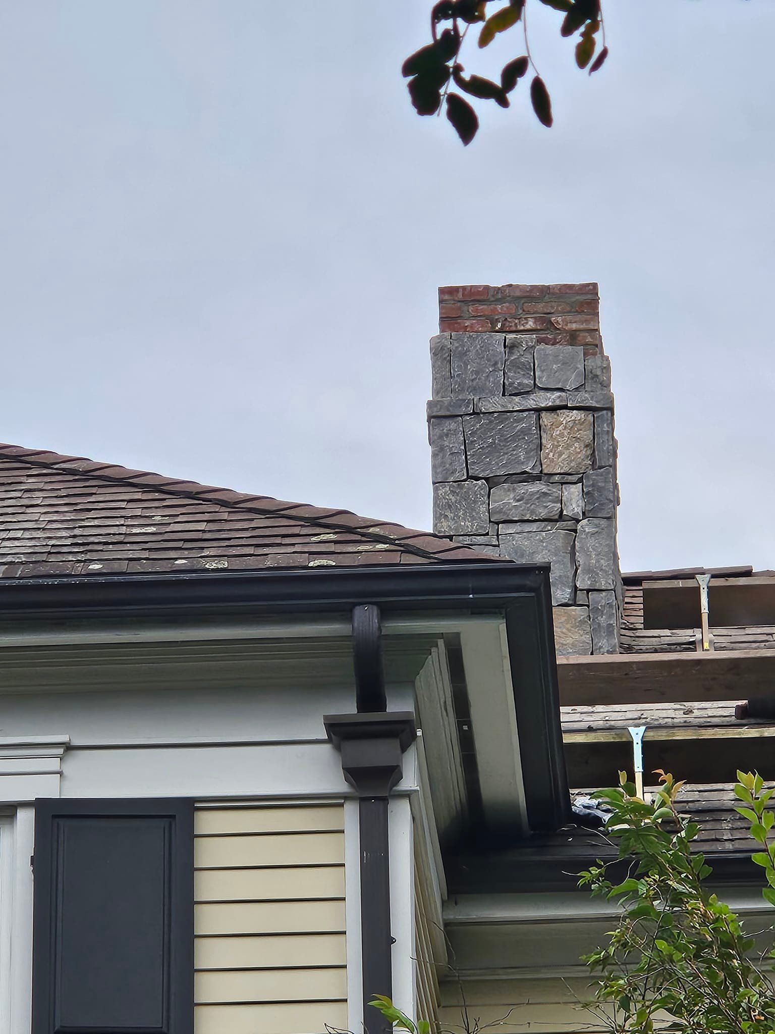 Chimney made of stone and brick on a house roof with a black gutter and window shutter.