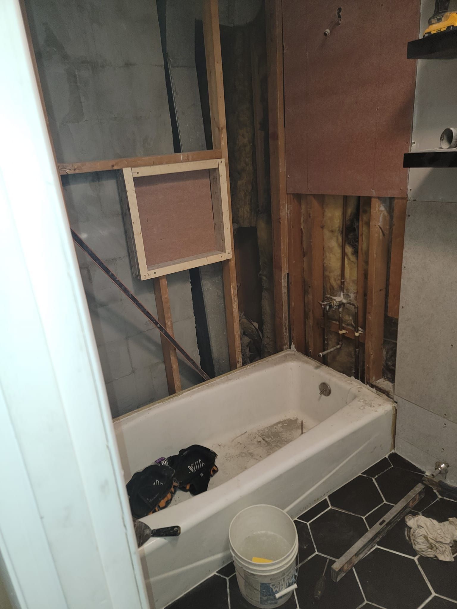 Bathroom renovation: tub, exposed studs, partially installed wall, a window-like frame, black and white tiled floor.