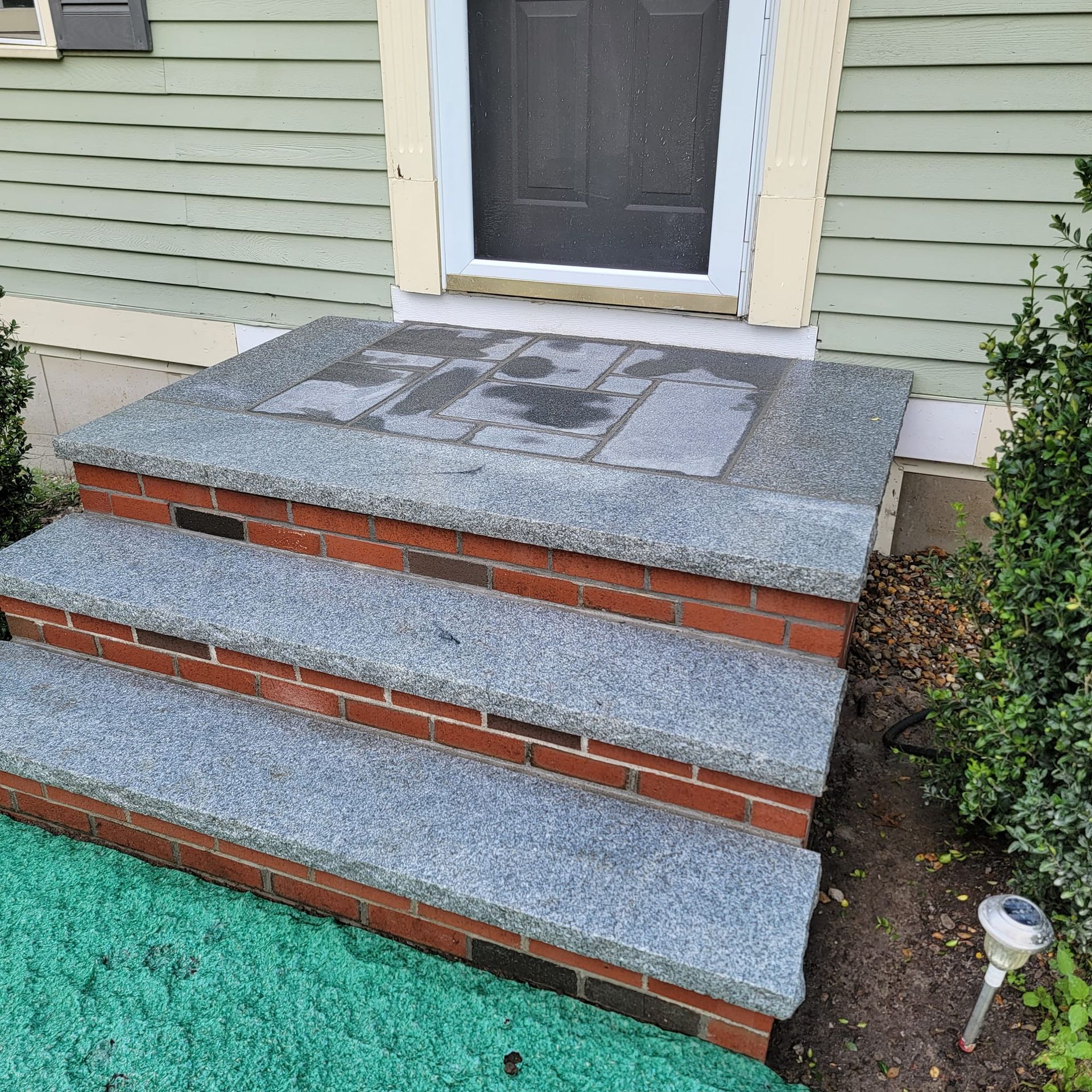 Brick steps with gray stone treads leading to a house entrance. Green siding and bushes flank the stairs.