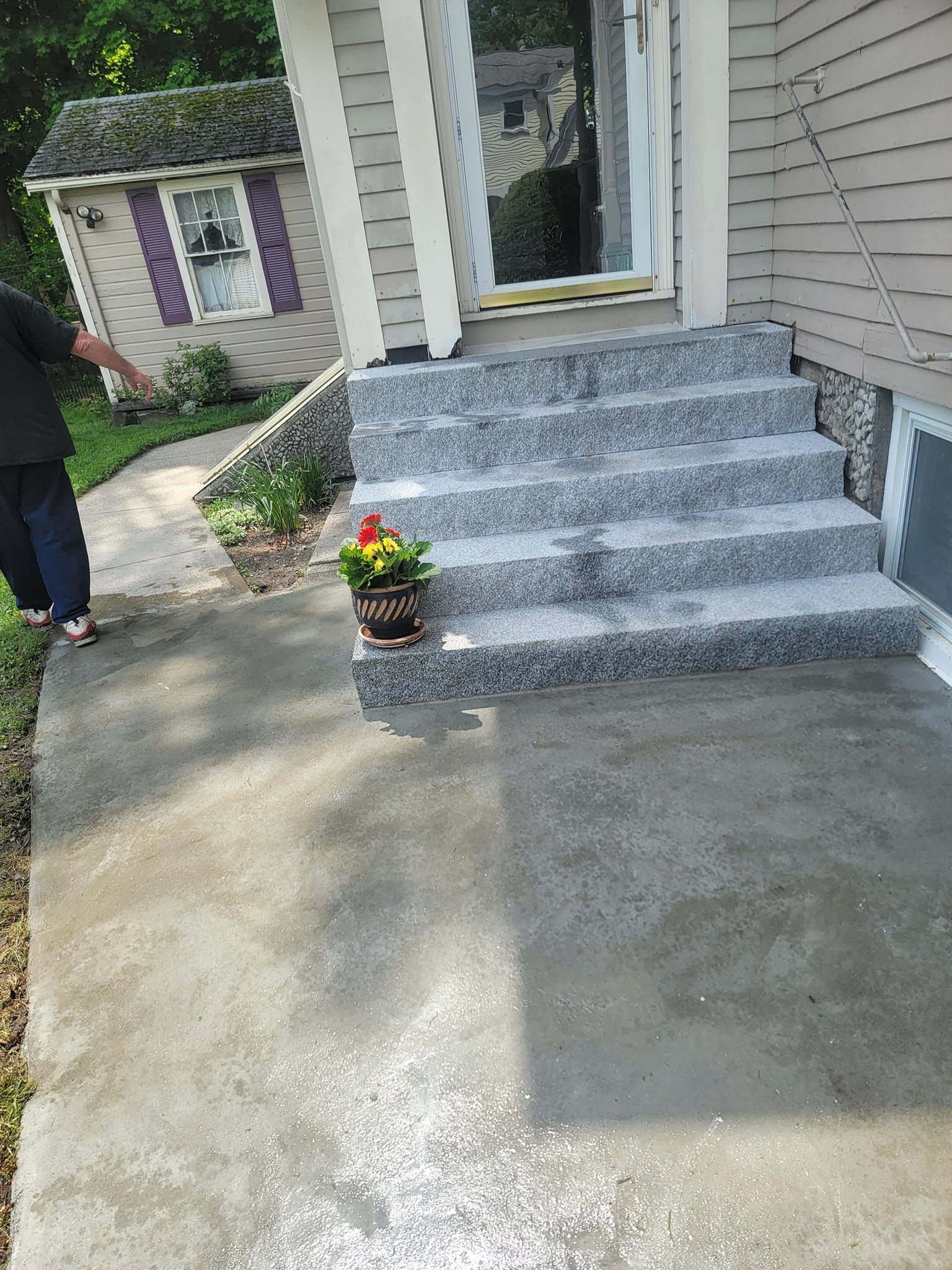 Concrete walkway and steps leading to a house entrance; person standing left; small purple building in the background.