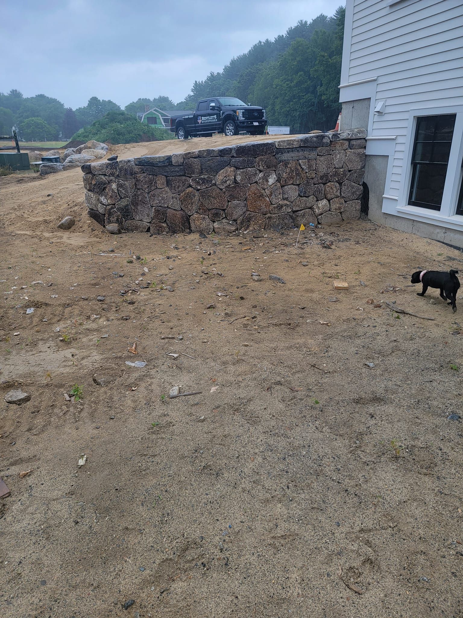 Stone wall next to a white house with a dirt yard and a black truck in the background, a small dog on the right.
