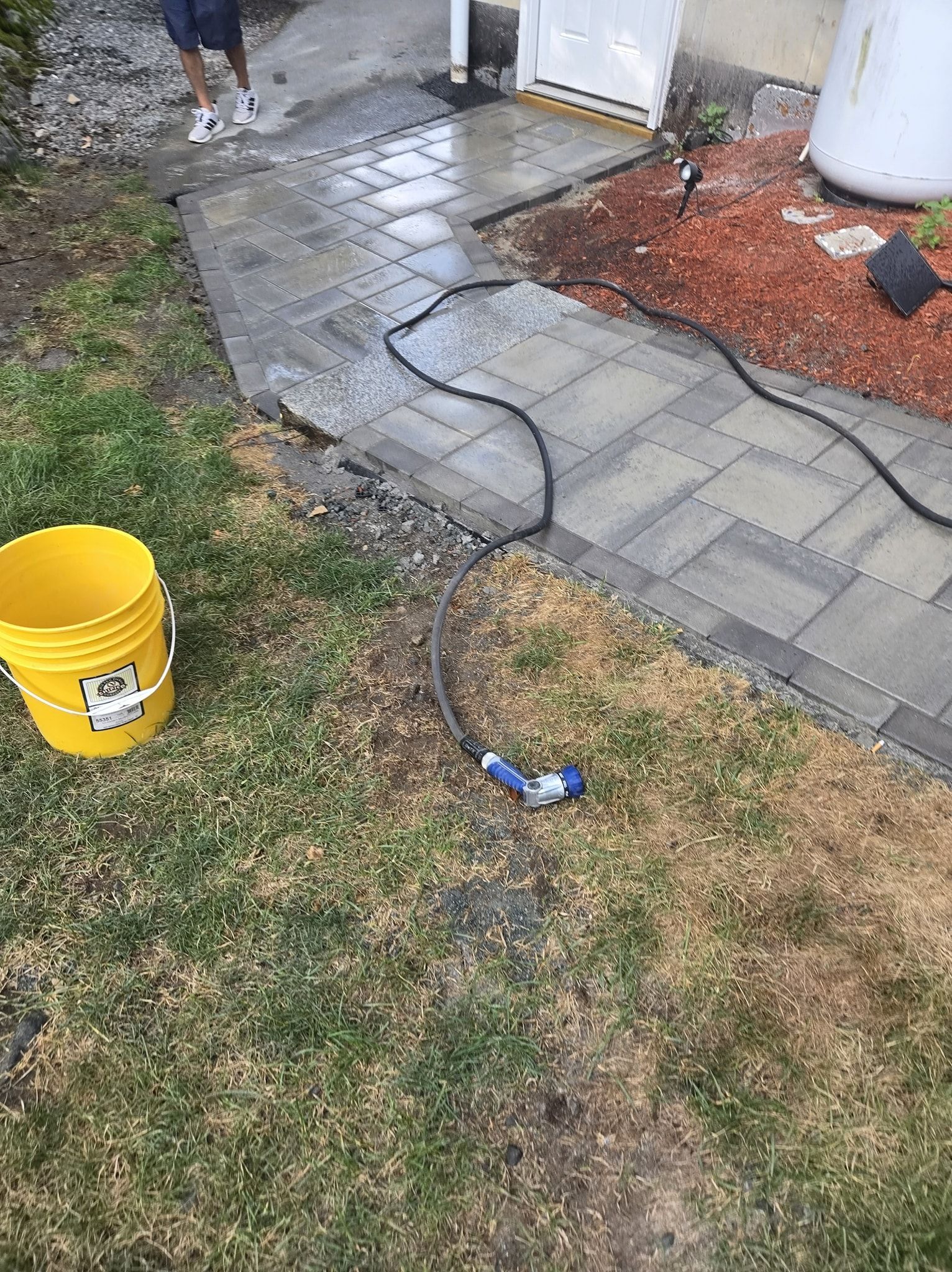 Person spraying water on new stone walkway with a yellow bucket nearby, on a sunny day.