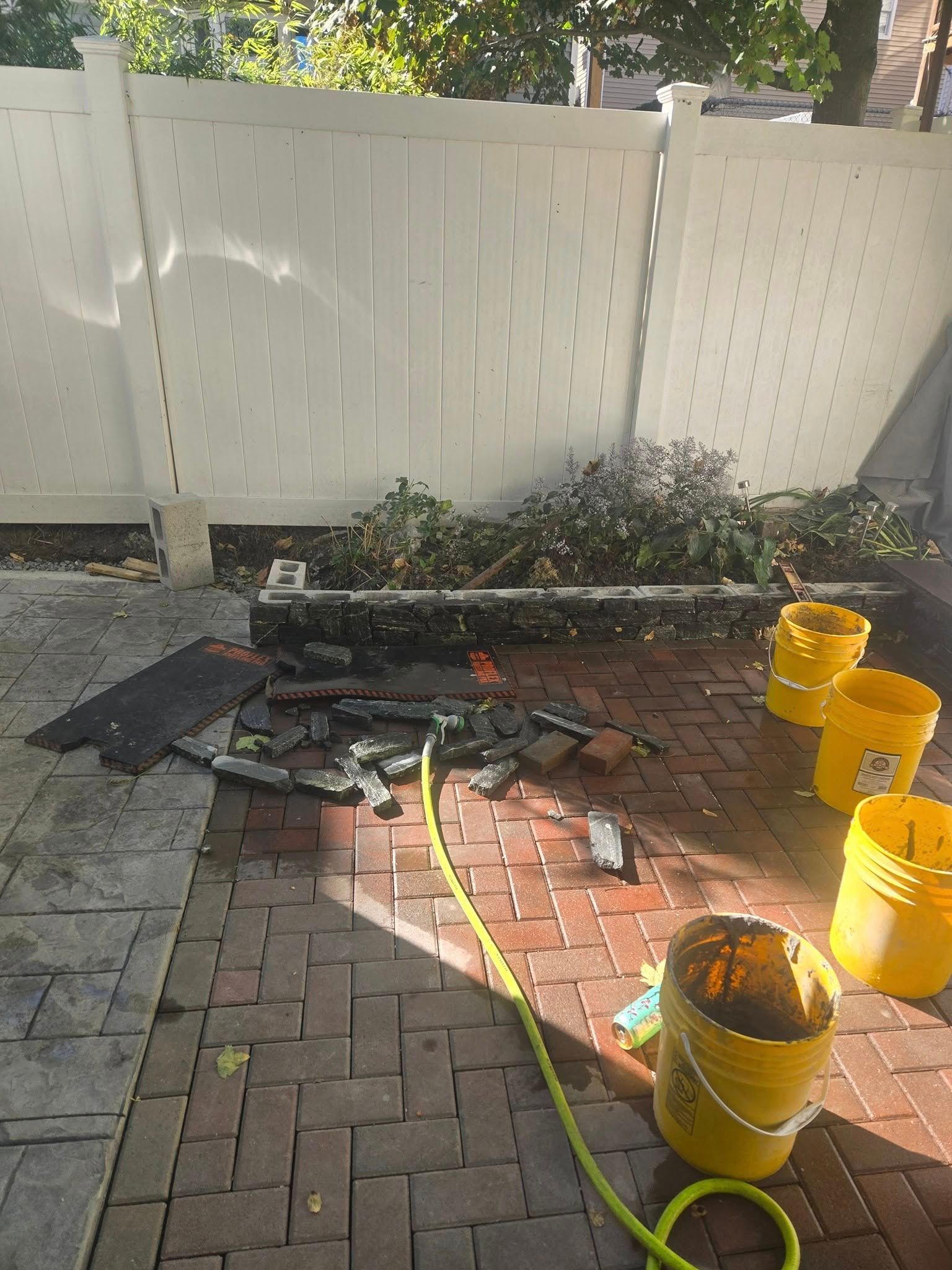 Patio with brick and concrete, debris, yellow buckets, hose, and white fence.