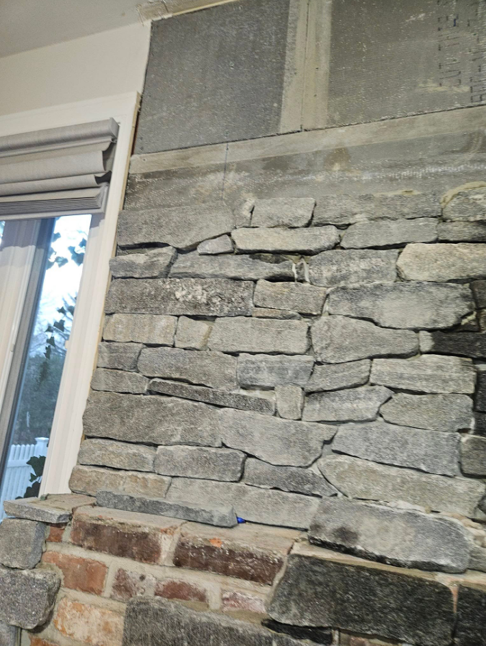 Stone veneer being applied to a fireplace wall. Gray stone, brick, and window visible.