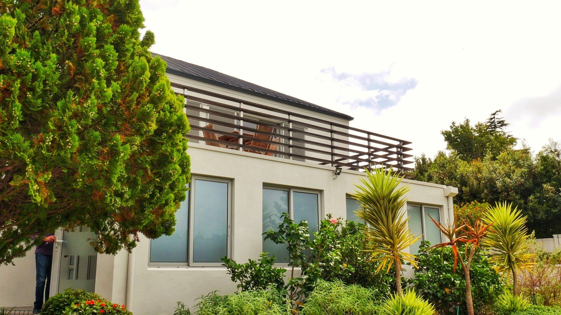 Een man staat voor een wit huis, omgeven door bomen en struiken.