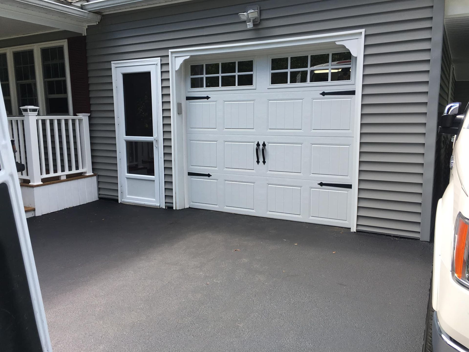 White garage door with black hardware, next to a screen door. Gray siding. Dark gray driveway.
