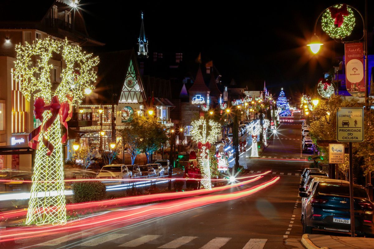 Rua da cidade à noite com luzes festivas de Natal, árvores iluminadas e rastros de luz dos carros que passam.