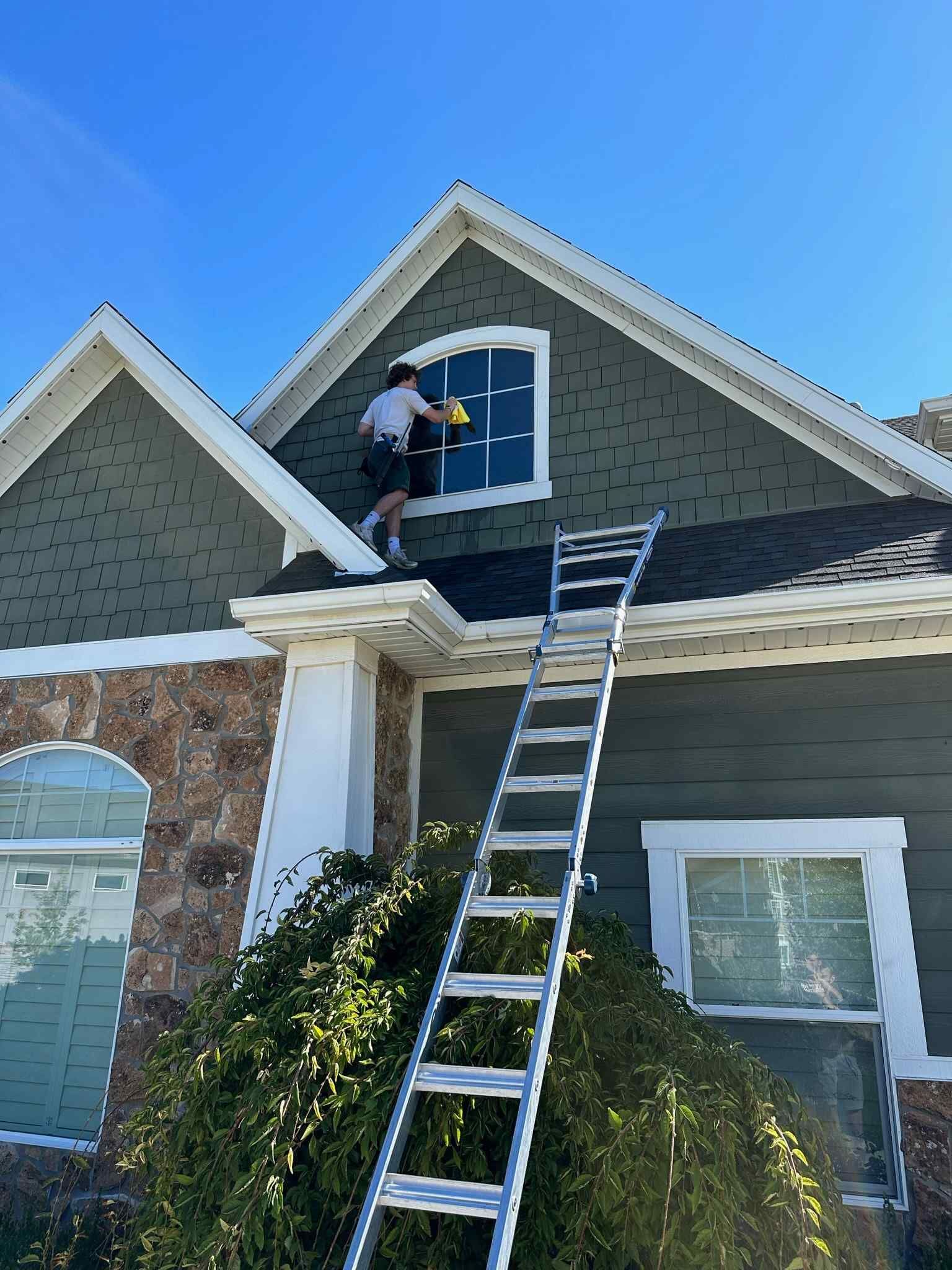 A man is standing on the roof of a house cleaning a window.