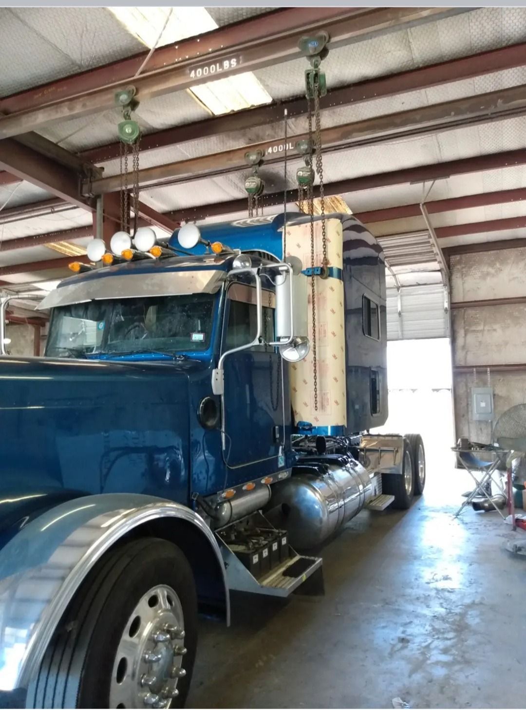 Blue semi-truck cab in a workshop, lifted by overhead pulleys. Silver fenders, chrome accents, and a custom cab extension.