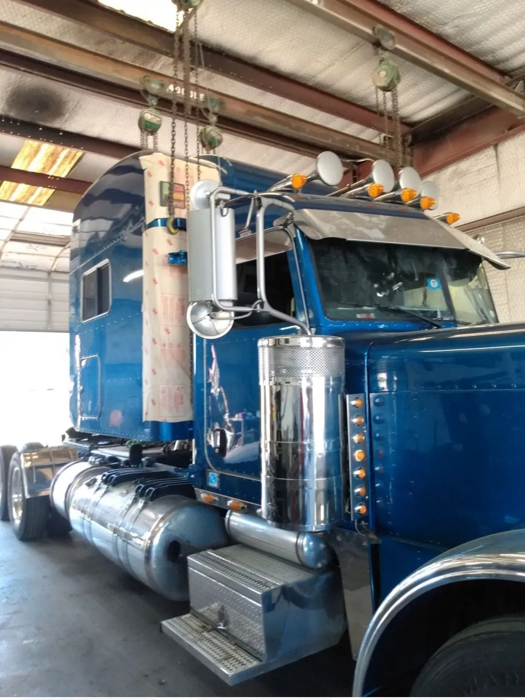 Blue semi-truck being worked on in a garage; a hoist is suspended above it.