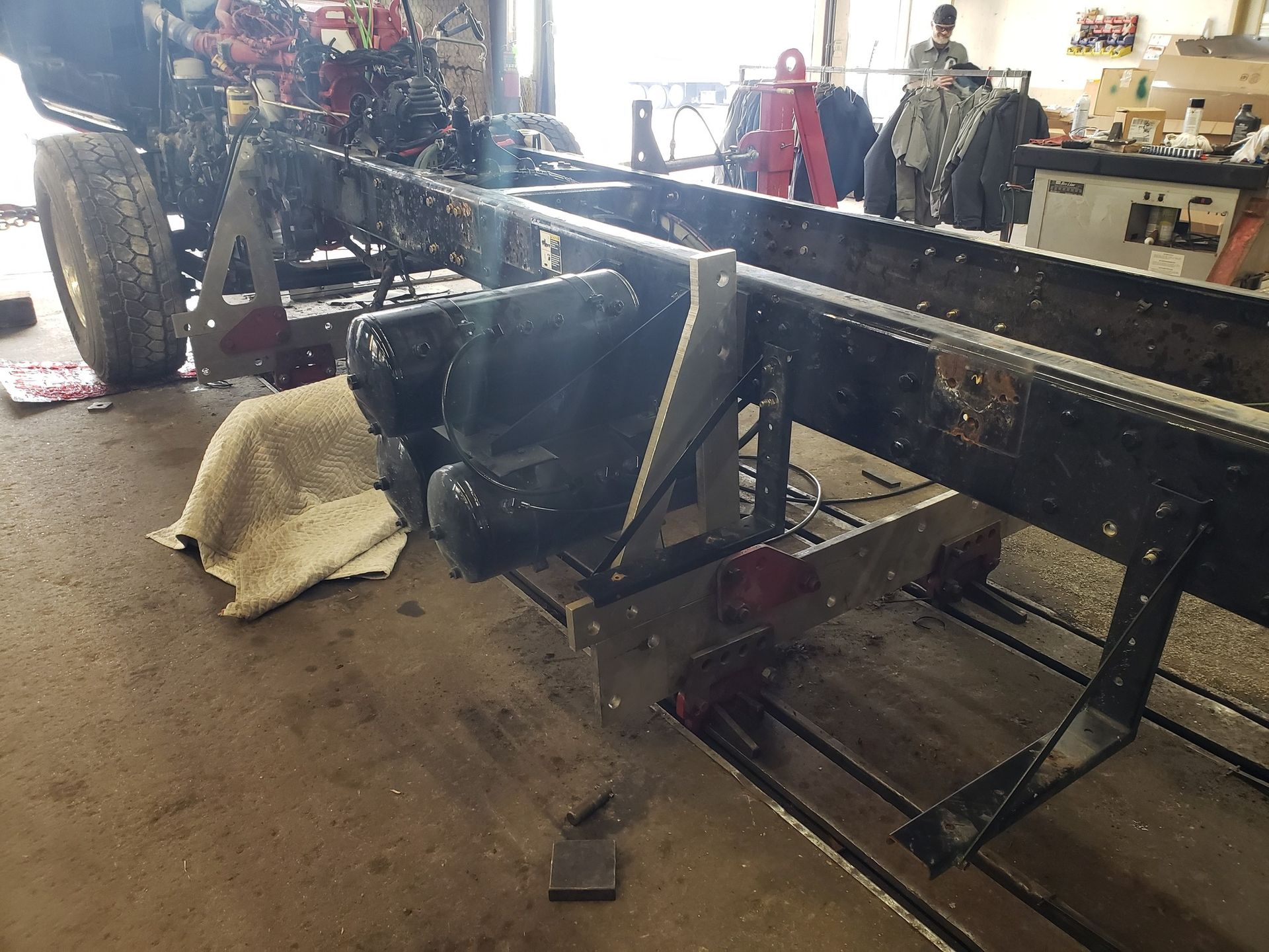 Truck chassis on jack stands in a shop. Dark metal, various parts, tools, and a person in the background.