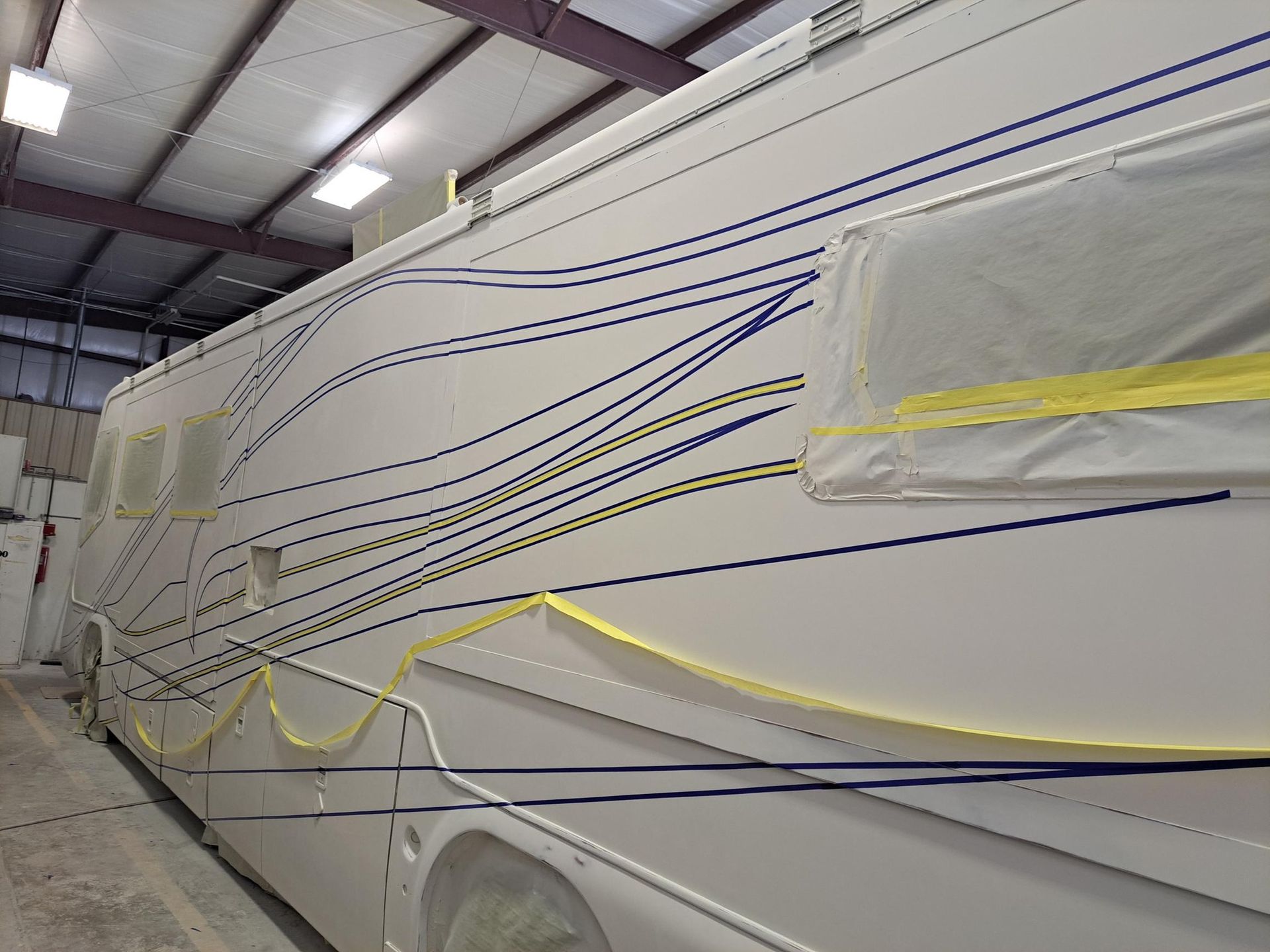 White RV being painted with blue and yellow design lines, in a workshop.