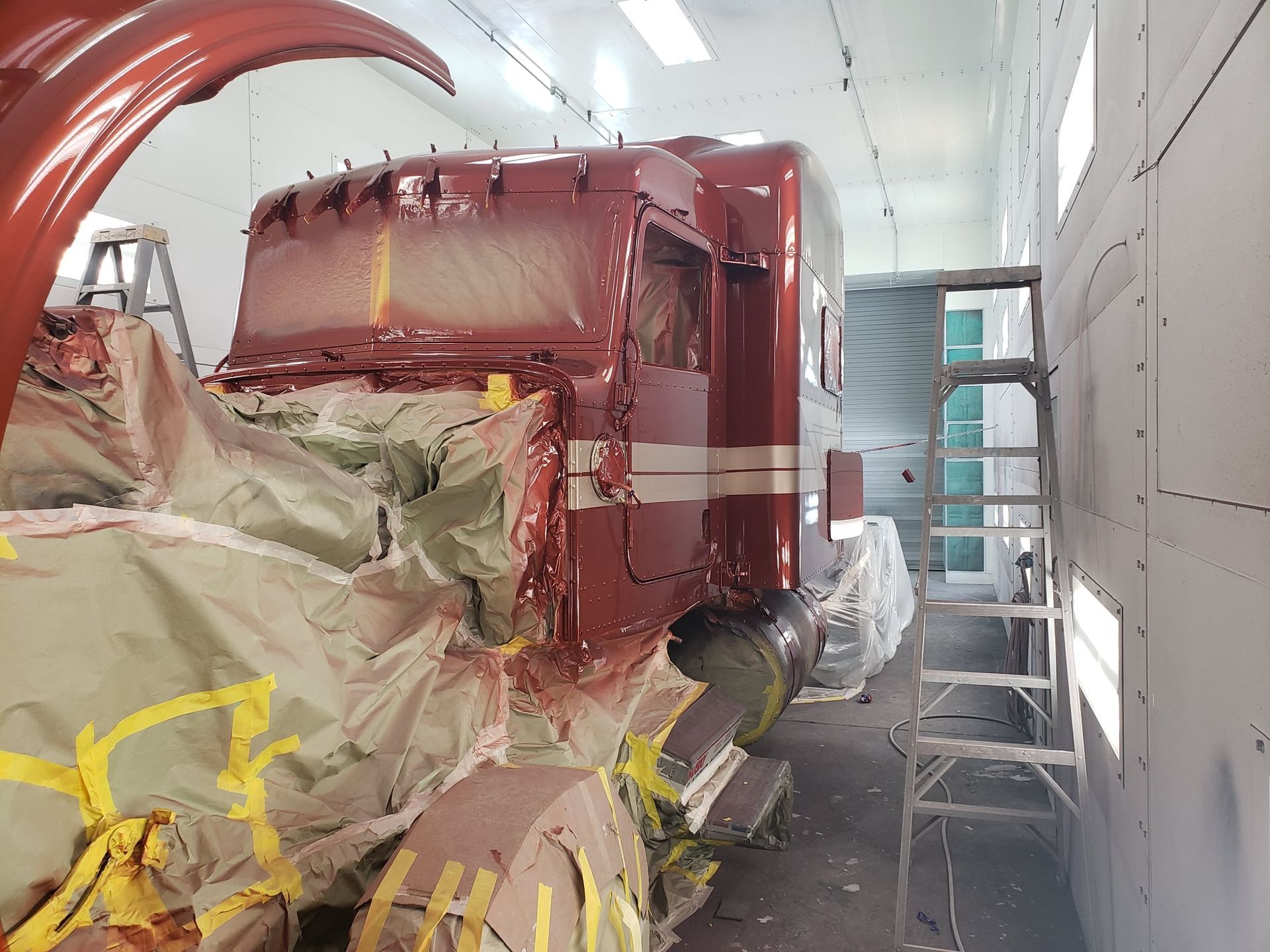 Truck cab in a paint booth, partially masked and sprayed with red paint; ladder nearby.