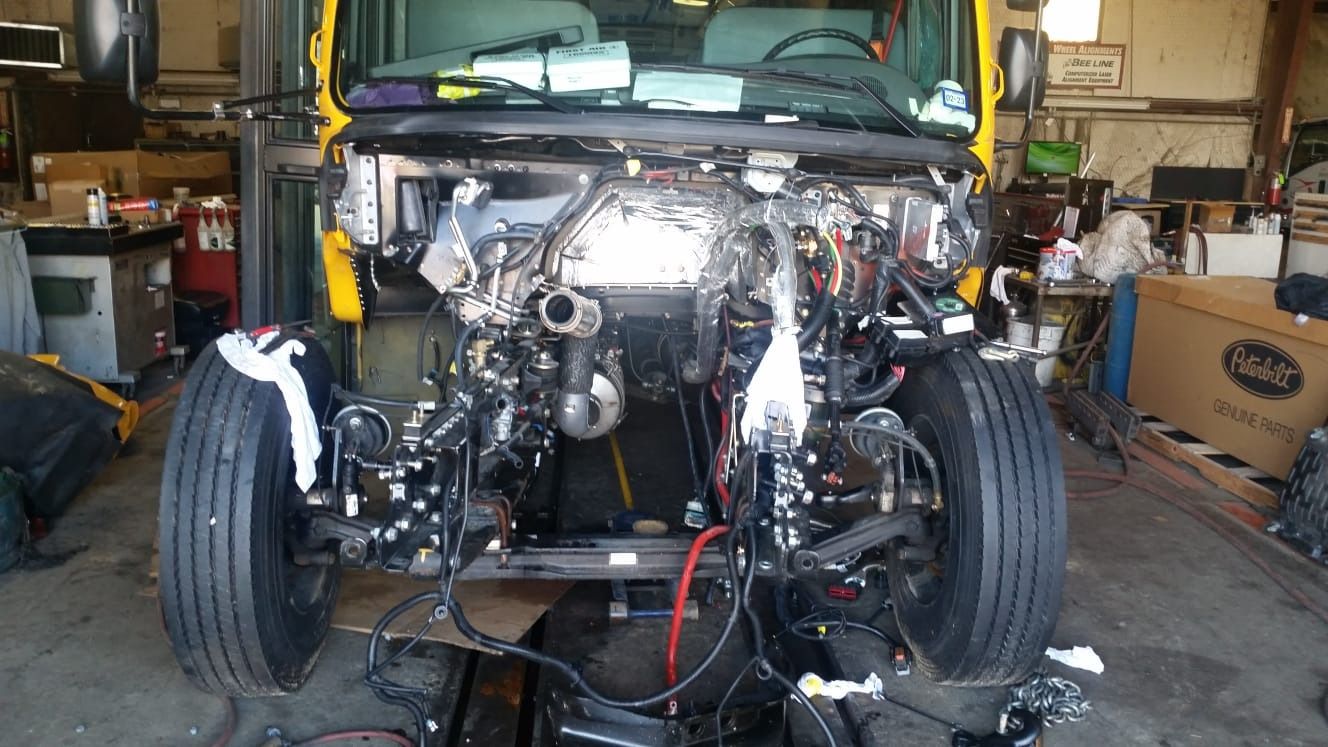 Front view of a yellow truck undergoing repair at a garage; the engine is exposed, and parts are removed.