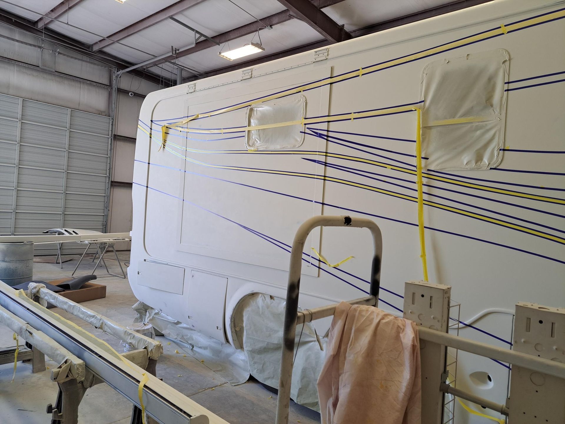A white RV in a workshop is prepped for painting. Windows are taped, with yellow and blue tape lines.