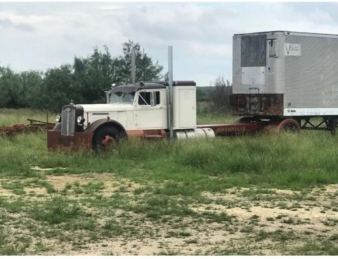 Old white and rust-colored semi-truck with trailer in a grassy field on a cloudy day.