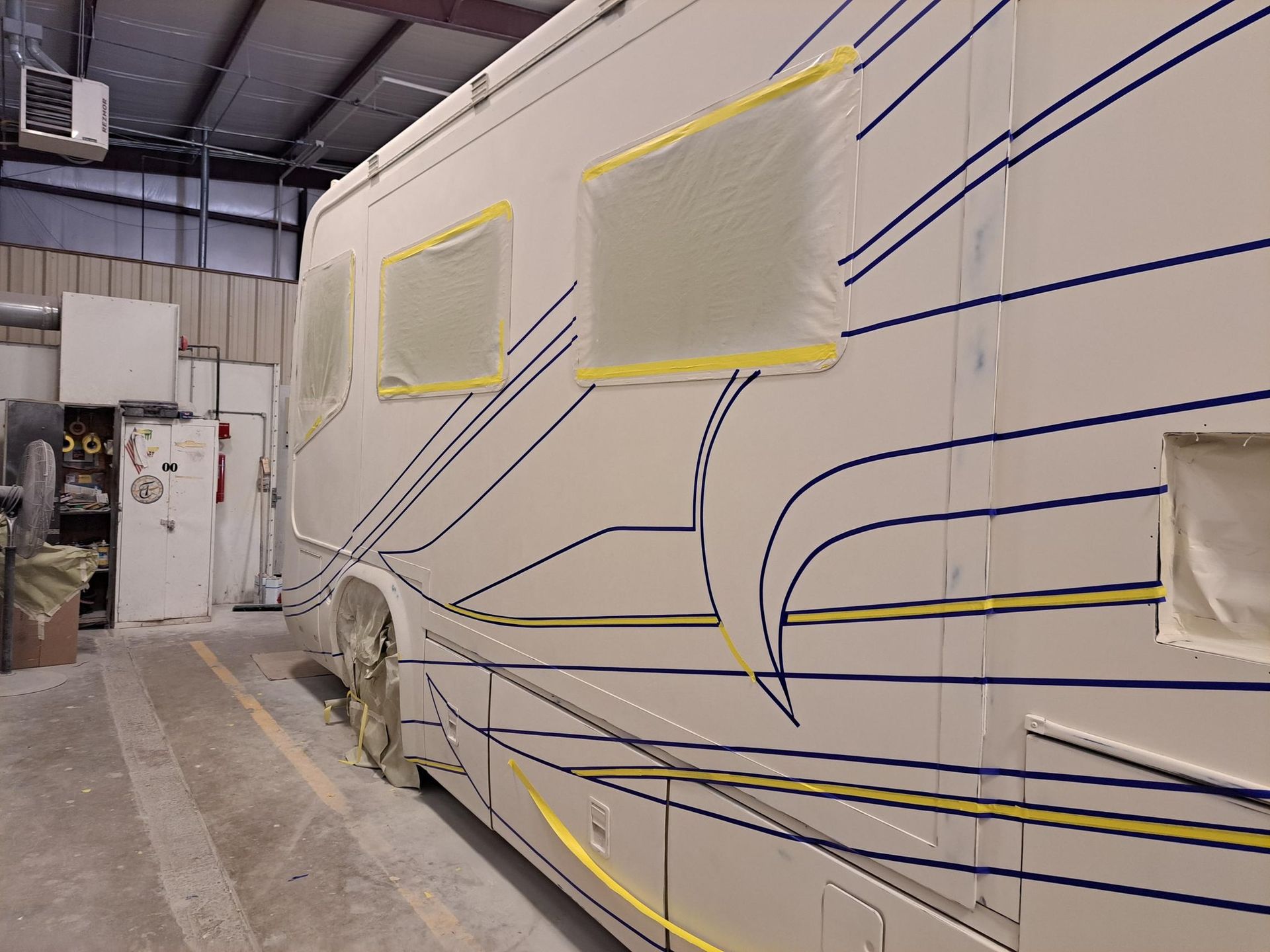 RV side with taped windows, blue and yellow design, in a workshop.