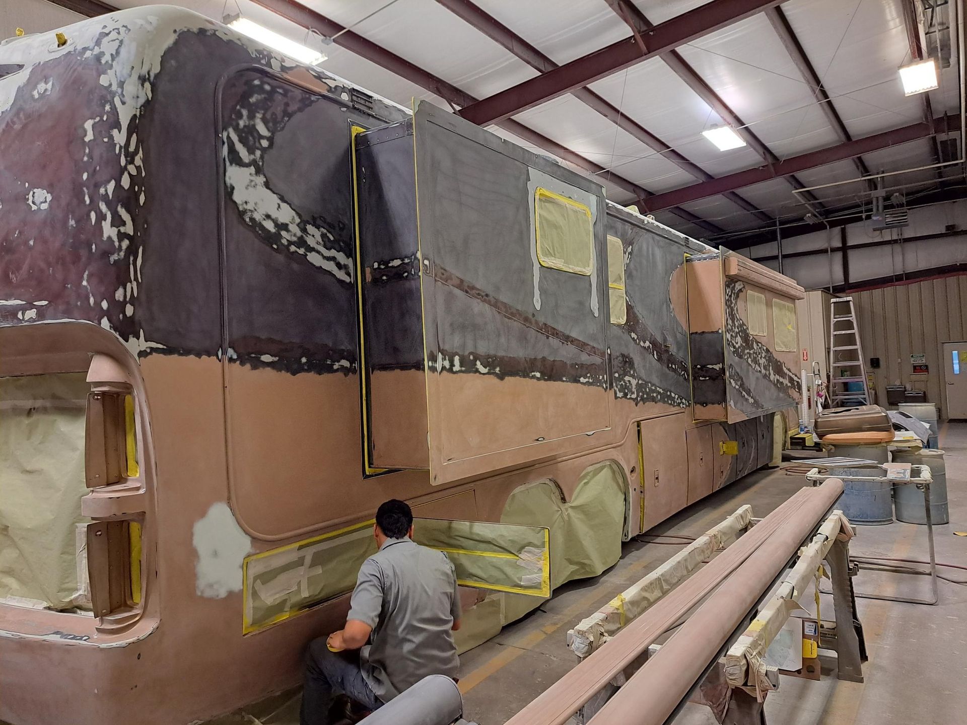 RV undergoing repair work in a workshop. A person is working on the exterior, applying filler.
