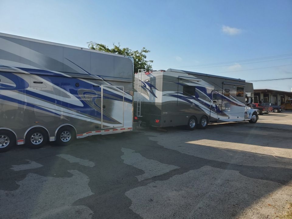 Two large, decorated trailers parked in a lot. One is a semi-truck with a silver and blue trailer. The other is a similar RV style trailer.
