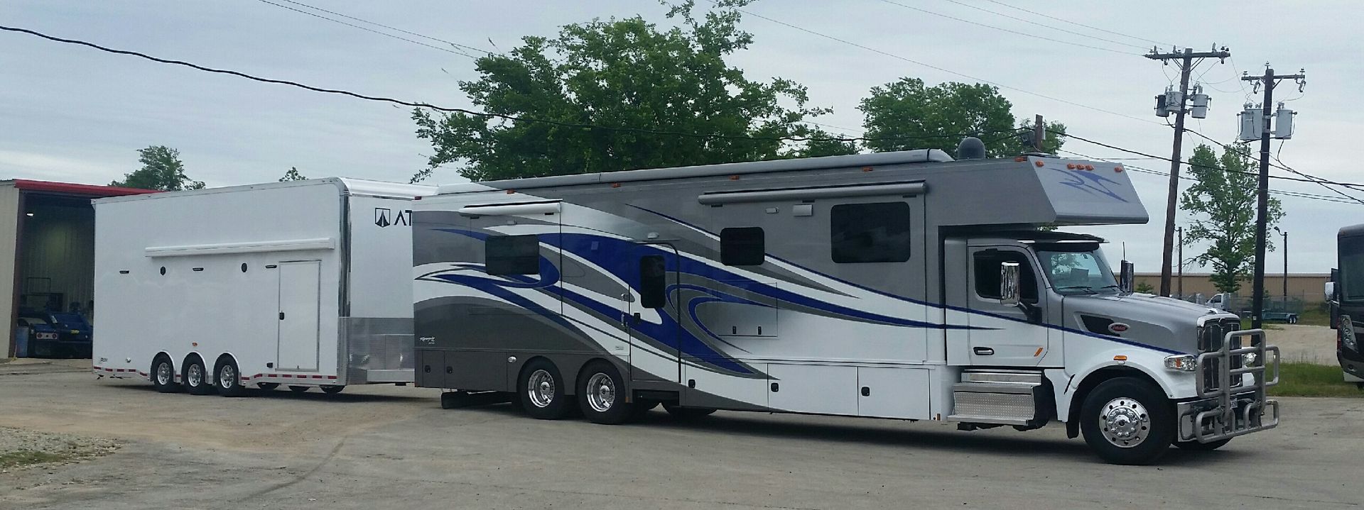 A large, custom truck and trailer parked outside. The rig is white, gray and blue. Trees and power lines in background.