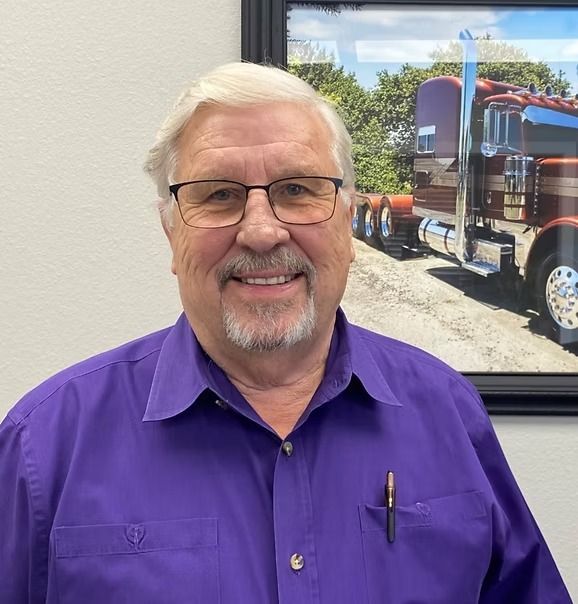 Man in purple shirt smiling, wearing glasses, in front of a photo of a truck.