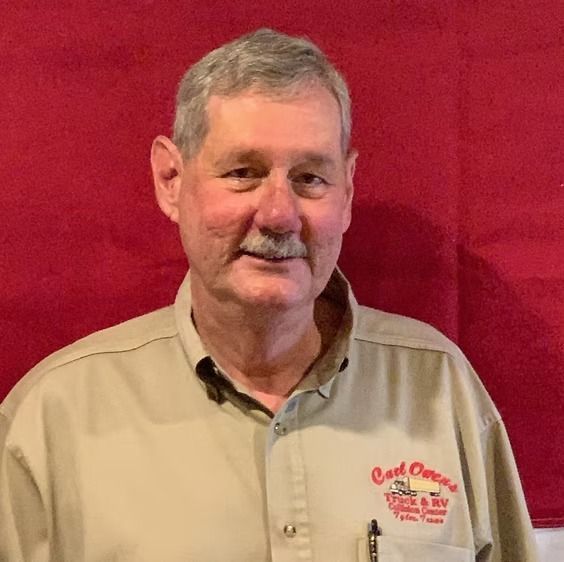 Man with gray hair and mustache, wearing tan button-down shirt with logo, in front of red backdrop.