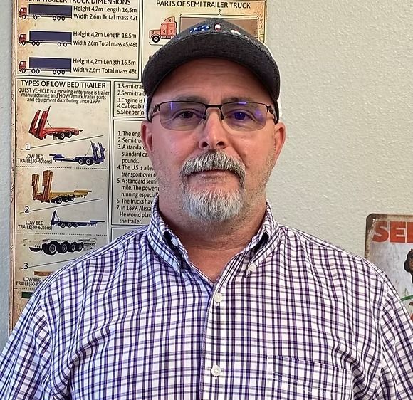 Man in cap and glasses, plaid shirt, in front of trucking info charts.