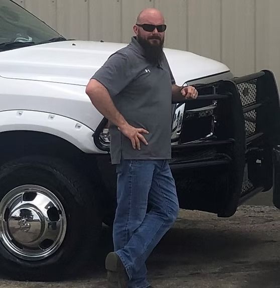 Man with beard wearing sunglasses, leans against white pickup truck with black grill guard.