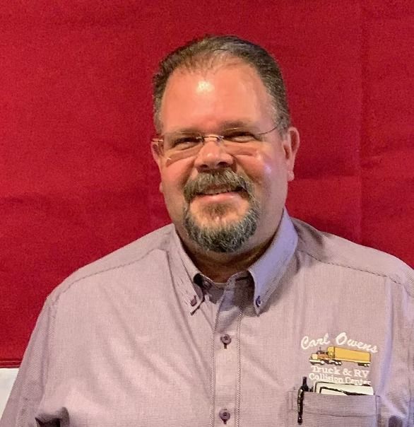 Man with glasses and goatee in a lavender shirt smiles, standing in front of a red backdrop.