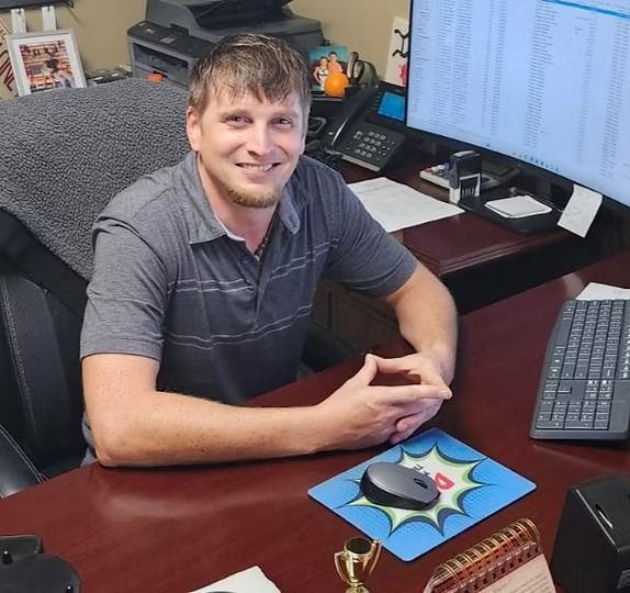 Man at desk, smiling. Gray shirt, hands clasped. Computer monitor, mousepad, keyboard visible. Office setting.