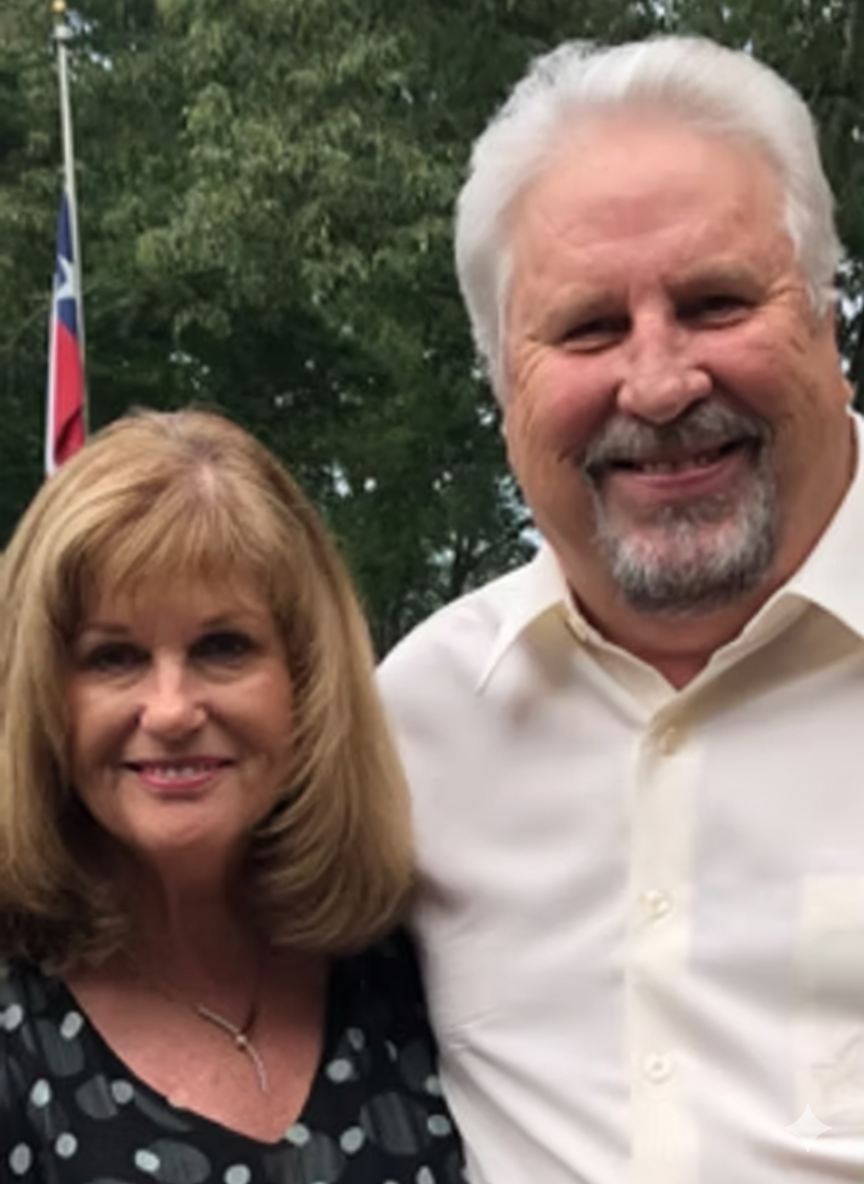 Couple smiling outdoors; woman with blonde hair, black top; man with white hair, button-up shirt; tree backdrop.
