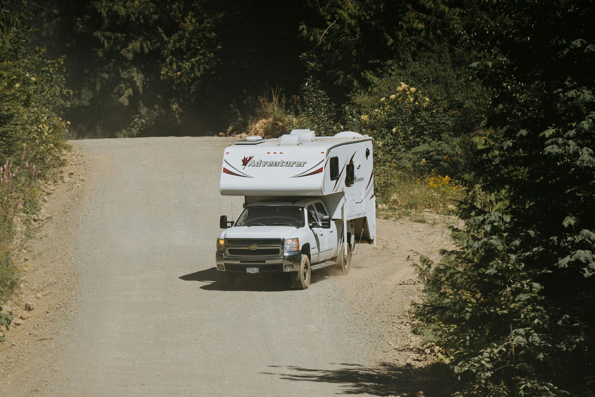 White truck with camper driving on a dirt road through a forest.