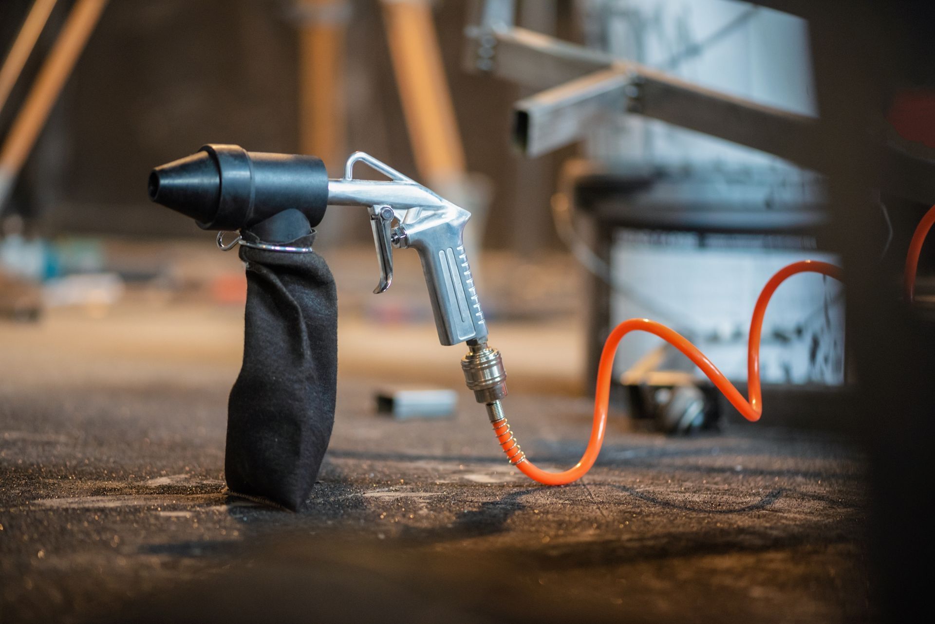Air sandblaster with black nozzle, silver gun, and orange hose, resting on a workshop floor.