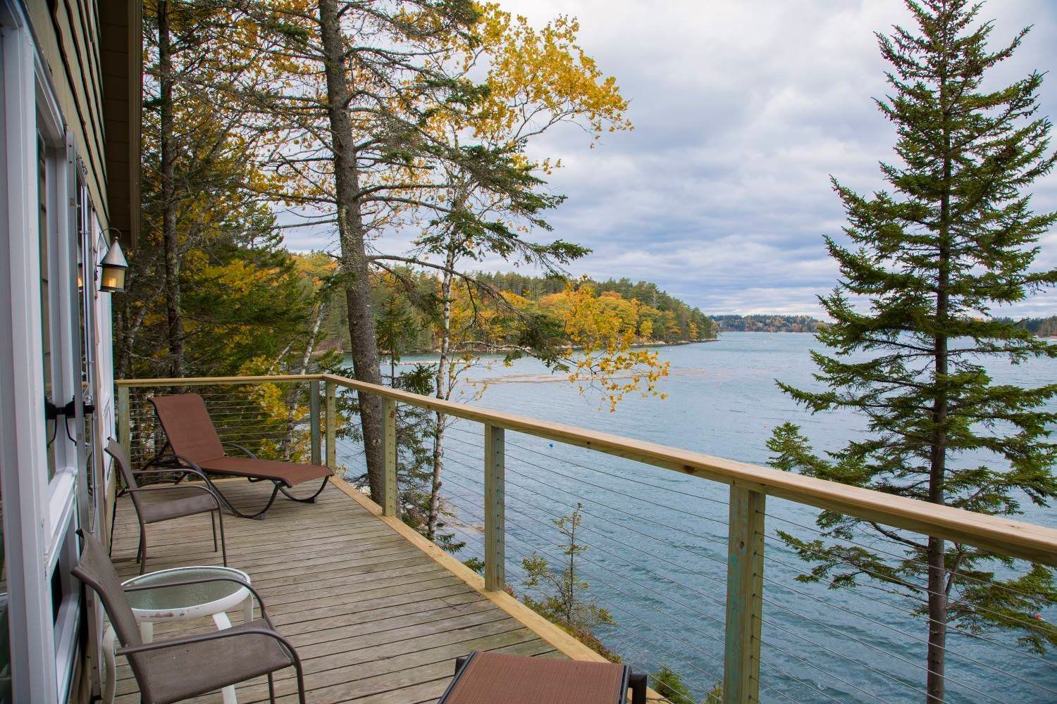 Deck overlooking a lake with two chairs, a small table, and trees. Cloudy sky above water.
