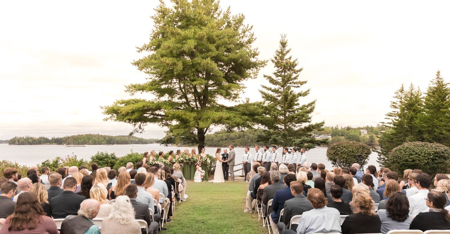 Wedding ceremony outdoors, overlooking water. Guests seated, bride and groom at altar under trees.
