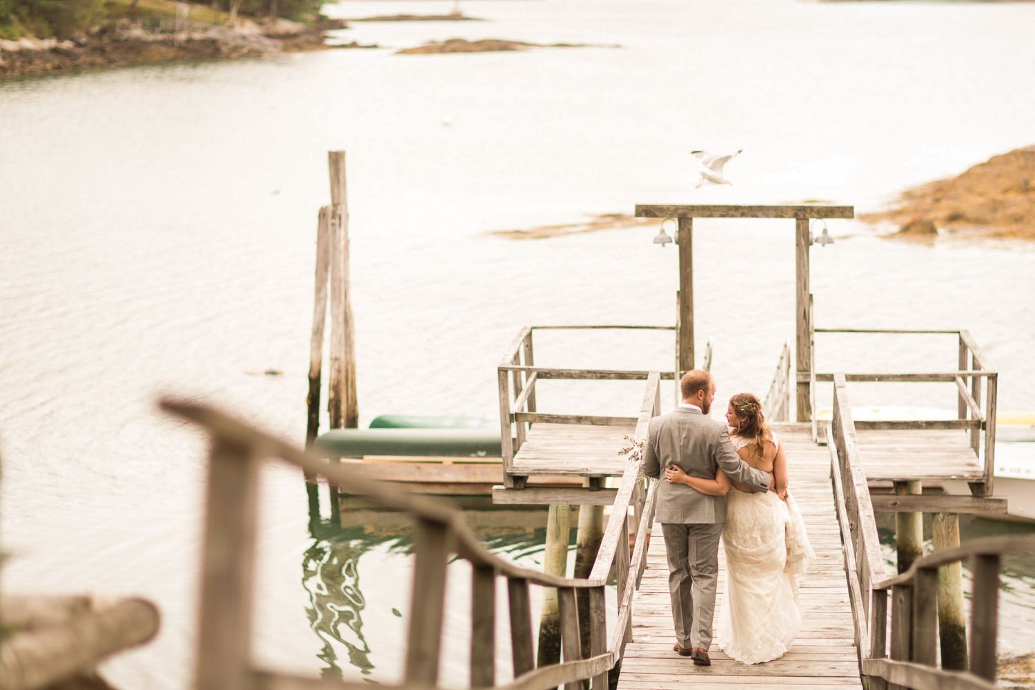 Couple in wedding attire walk arm-in-arm down a wooden dock towards the water.
