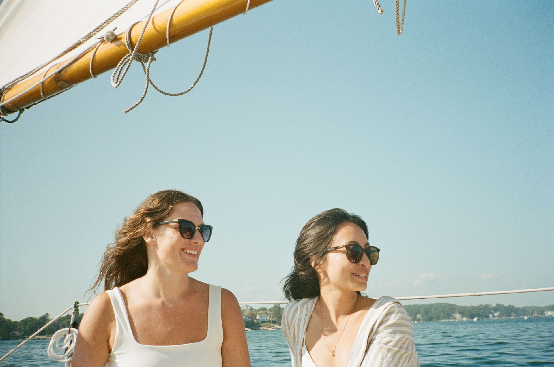 Two women wearing sunglasses smile on a sailboat under a blue sky.