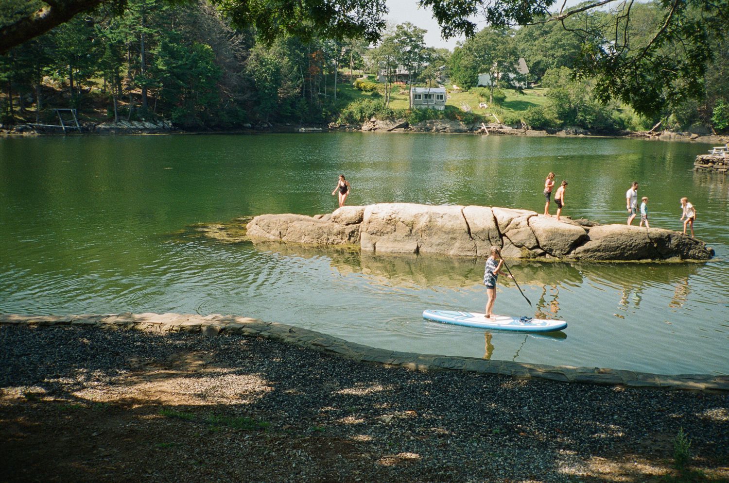 People stand on a rocky island in a green lake. One person paddles a paddleboard. Trees line the shore.