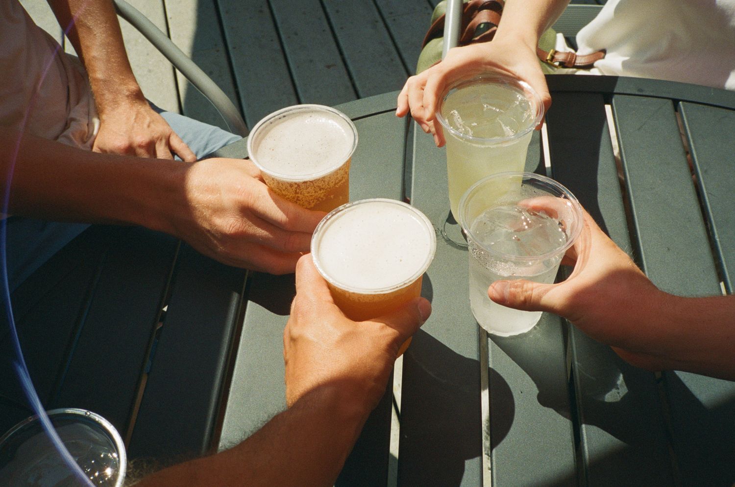 Four people toasting drinks outdoors. Beer and cocktails in plastic cups on a table.