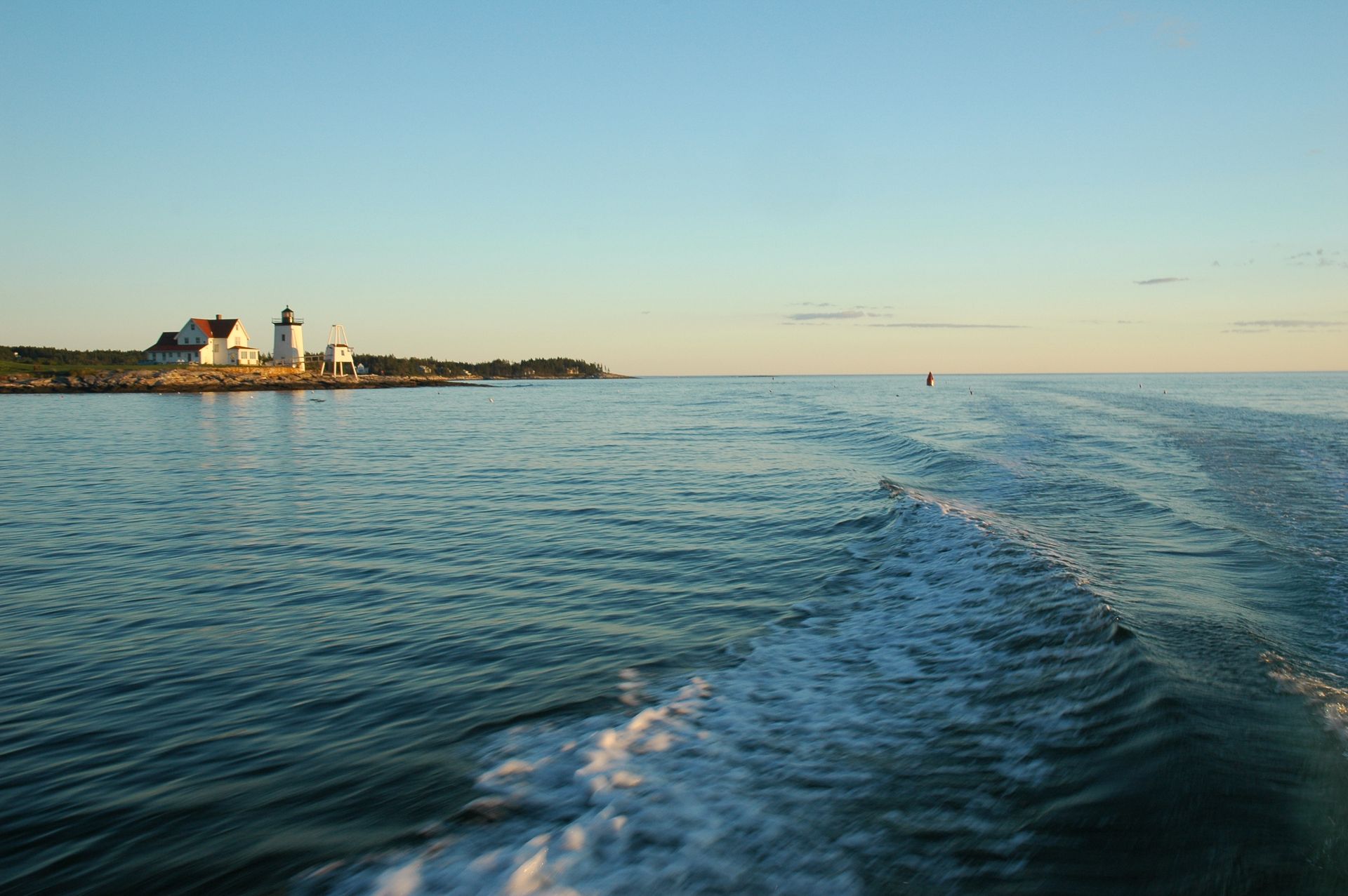 Ocean view with lighthouse on shore, boat wake in the foreground, sunny day.