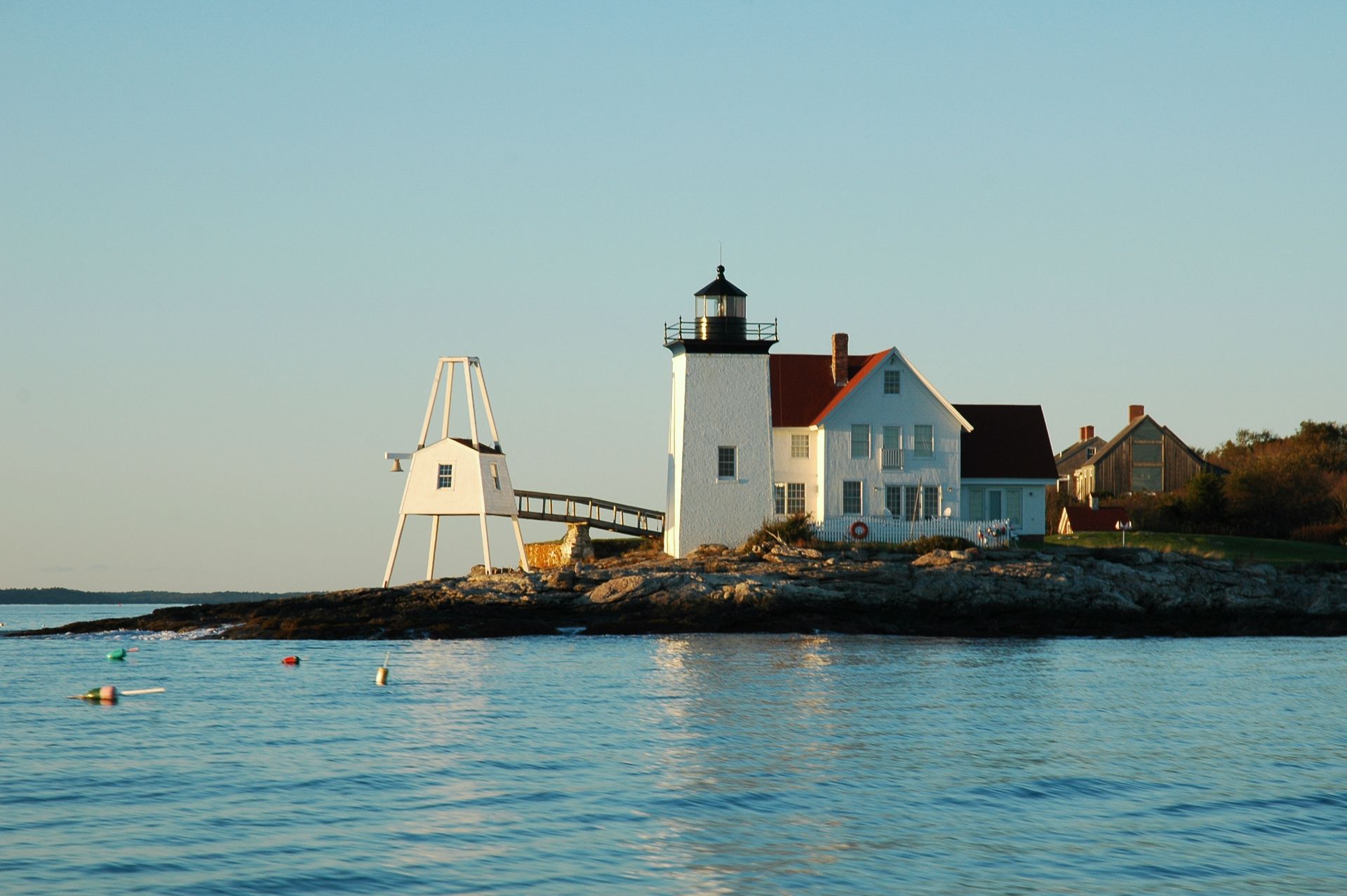 White lighthouse with red roof on rocky shore, small white square structure nearby, blue water and sky.