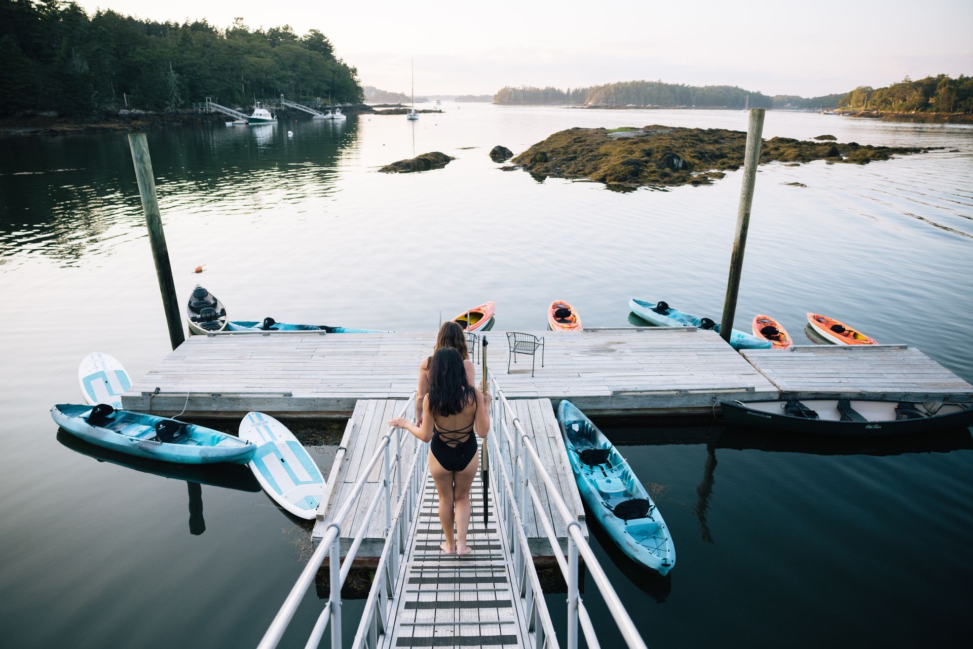 Woman in a swimsuit walks towards kayaks on a wooden dock at a calm, coastal inlet.