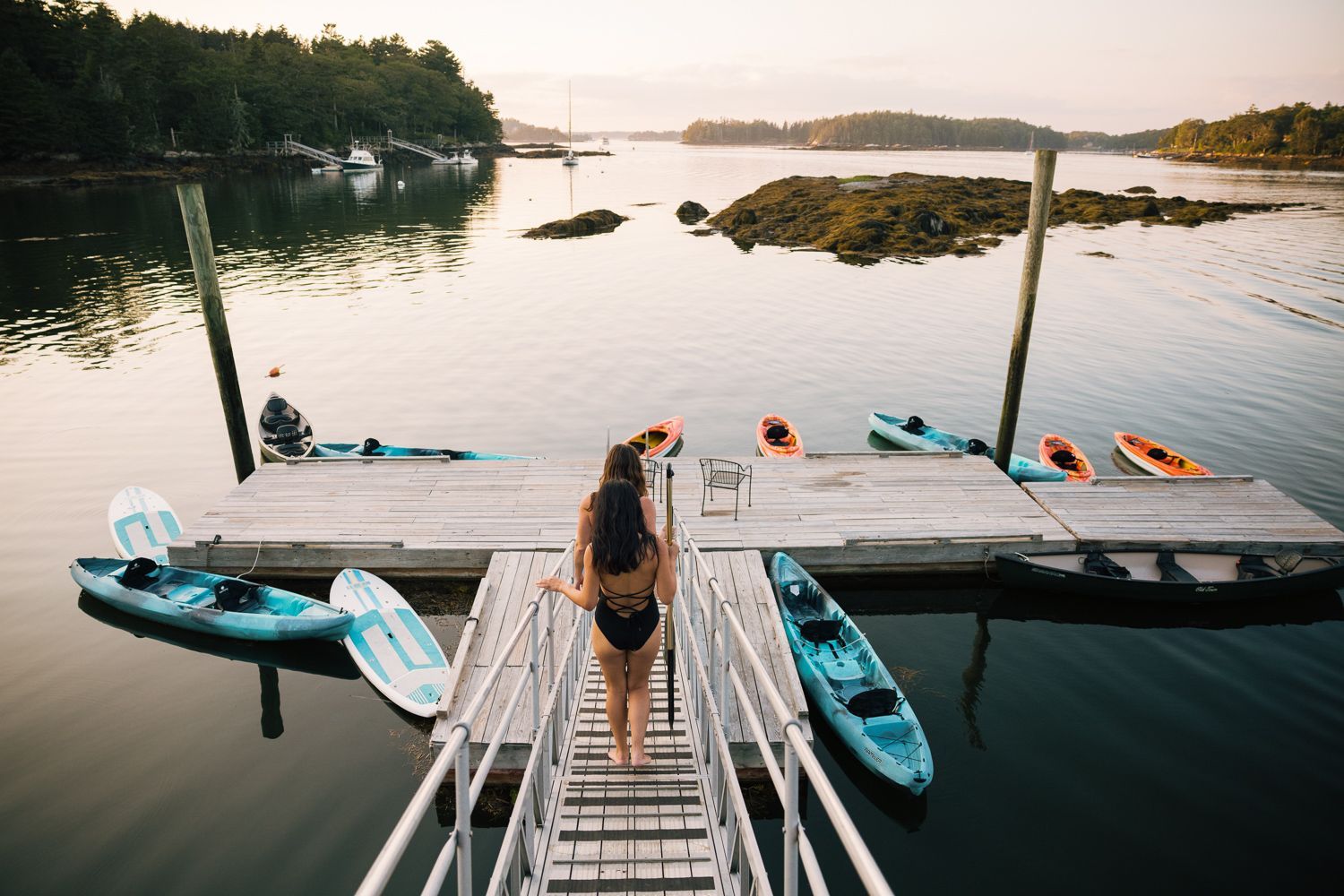 Woman in swimsuit walks towards kayaks on a dock. Calm water, trees in the background.