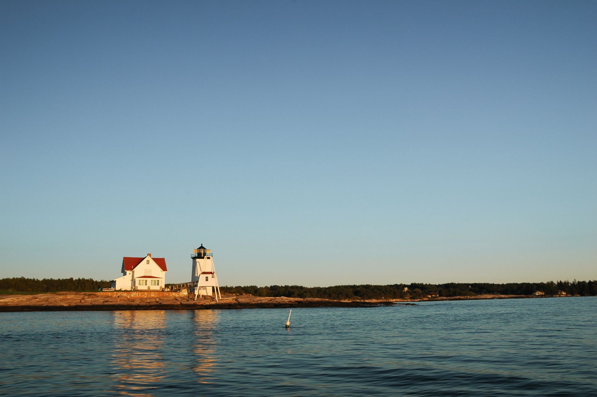 Lighthouse and adjacent building on a rocky shore, clear blue sky, calm water.