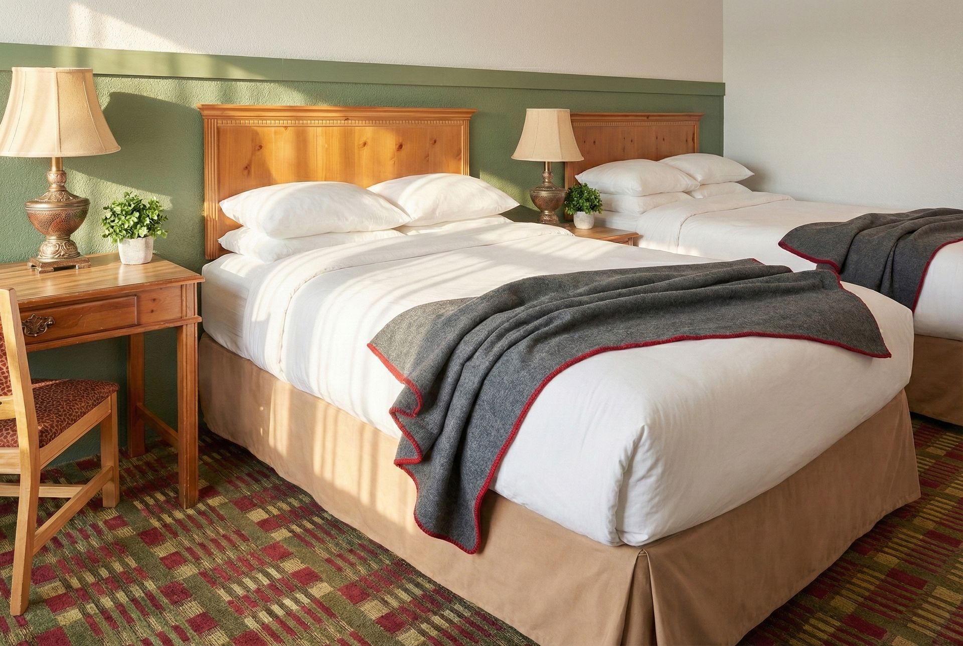 Two hotel beds with white linens, wooden headboards, and a blanket. A wooden desk and lamp are on the left.